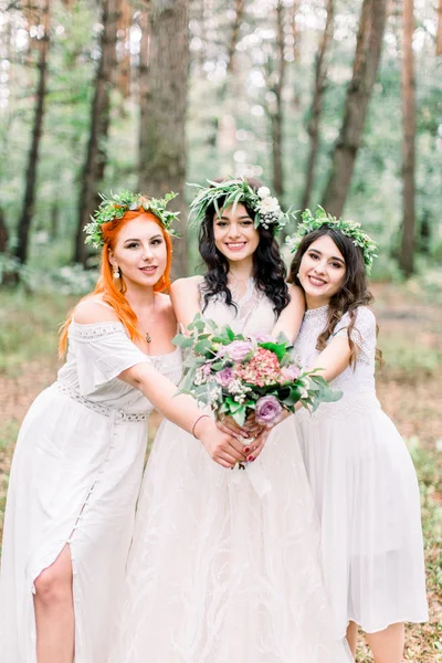 Feliz novia sonriente posando con sus damas de honor en vestidos blancos al aire libre en el bosque. Novia y damas de honor en corona floral en cabezas con ramo de boda rústico . —  Fotos de Stock