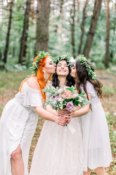 Dos hermosas damas de honor besan las mejillas de las novias mientras están en el bosque. Chicas con coronas florales y vestidos rústicos posando en el bosque —  Fotos de Stock