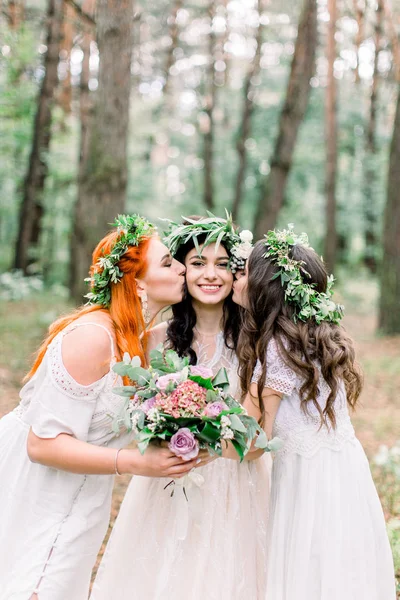 Dos hermosas damas de honor besan las mejillas de las novias mientras están en el bosque. Chicas con coronas florales y vestidos rústicos posando en el bosque —  Fotos de Stock