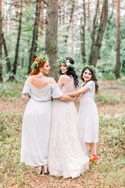 Vista posterior de la novia bastante alegre y damas de honor en vestidos blancos y coronas flotantes divertirse en el día de la boda, caminando en el bosque de pinos . —  Fotos de Stock
