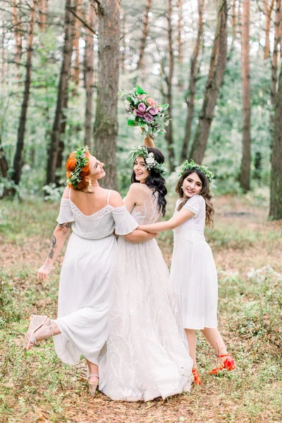 Deux jolies demoiselles d'honneur entourent la mariée avec le dos tourné, portrait complet sur fond de forêt. Femmes en robes blanches et avec des couronnes florales sur la tête — Photo