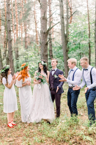 Невеста и жених с подружками невесты и шафер веселятся и улыбаются стоя на открытом воздухе в зеленом парке или лесу — стоковое фото