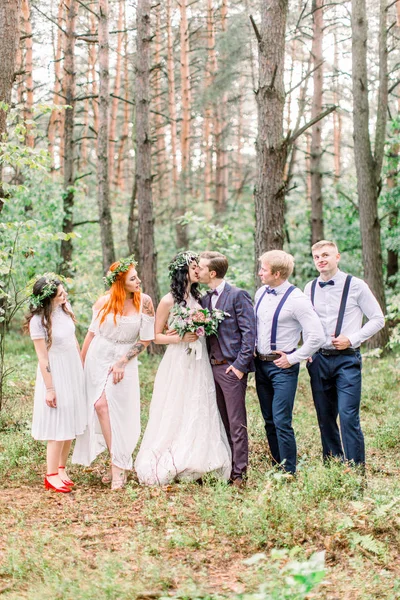 Piękne całowanie pary nowożeńców z przyjaciółmi bawiących się razem. Szczęśliwe druhny i druhny z panną młodą i panem młodym na spacerze po lesie — Zdjęcie stockowe