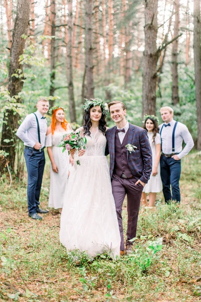 Šťastný úsměv novomanželé nosí elegantní svatební šaty v rustikálním stylu a jejich freinds stojí bok po boku v borovém lese — Stock fotografie