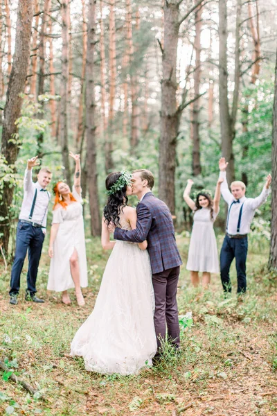 Stylish rustic wedding couple with bridesmaids and groomsmen at forest on wedding day. Back view of groom and bride, embracing and kissing. Their friends smiling and holding their hands on the air. — Stock Photo, Image