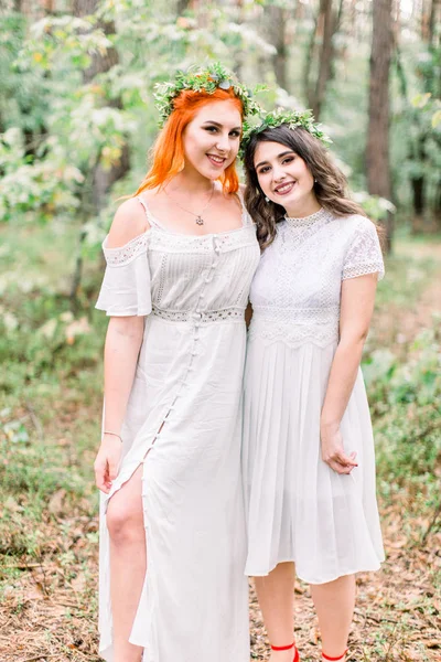 Belle deux filles demoiselles d'honneur en robes blanches et des couronnes florales sur la tête posant et regardant à la caméra le jour du mariage, fond de forêt — Photo
