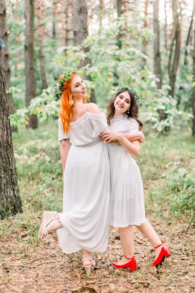 Drôle deux belles filles demoiselles d'honneur dansent dans la forêt. Deux demoiselles d'honneur en robes blanches et des couronnes sur les têtes souriant et s'amusant ensemble — Photo