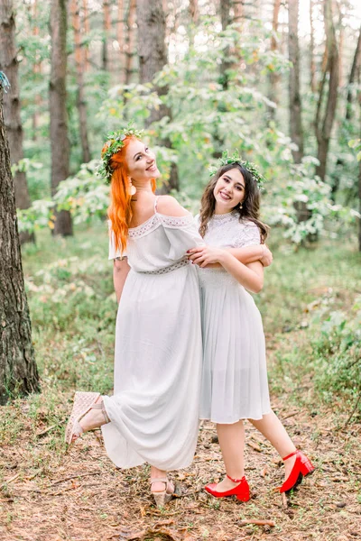 Drôle deux belles filles demoiselles d'honneur dansent dans la forêt. Deux demoiselles d'honneur en robes blanches et des couronnes sur les têtes souriant et s'amusant ensemble — Photo