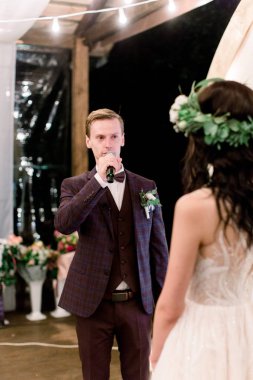 Düğün günü. Takım elbiseli çekici bir damat, bir yemin ederken, gelinine zarif bir kırsal elbise içinde ve başında çelenk ile gece düğün töreninde bakıyor. Açık havada ahşap çadır restoranı.
