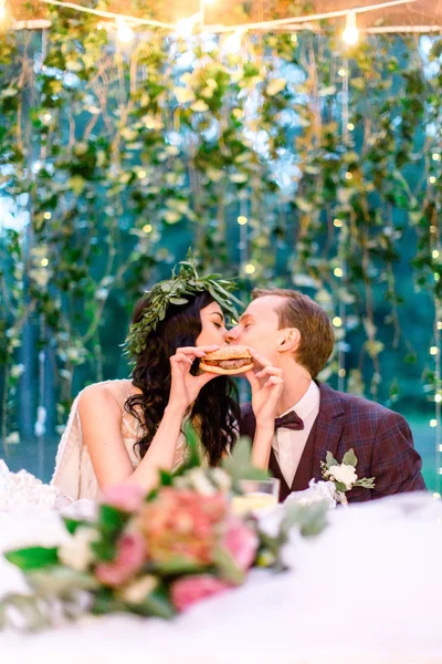 Stylish wedding couple eating fastfood and kissing, bride holds tasty burger in hands. Wedding outdoors — 스톡 사진