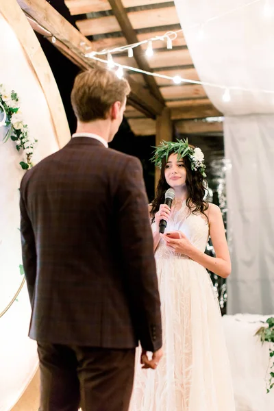 Pretty bride in wedding dress and floral wreath on head is speaking an oath to her groom during night wedding ceremony, standing near the rustic arch. Outdoors. — 스톡 사진