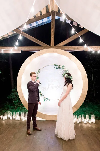 Night wedding ceremony outdoors. Bride and groom swear an oath each other on wedding arch background. Beautiful just married couple. Rustic wooden restaurant with light bulbs