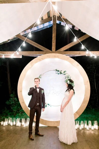 Wedding ceremony in the open air in the stylish rustic wooden tent in forest. Wooden arch with flowers and greenery. The bride and groom are exchanging oaths. — Stock Photo, Image