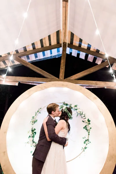 Young handsome groom in dark suit and bride in white dress and floral wreath standing near arch and kissing. Happy couple on night wedding ceremony in rustic restaurant outdoors — 스톡 사진