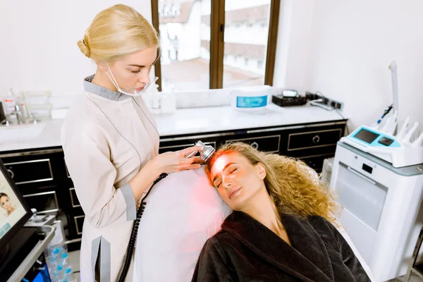 Kadın doktor yüz derisini tazelemek için prosedür uyguluyor. Güzel sarışın kadın yüz bakımı görüyor, klinikteki pigmentasyonu kaldırıyor. Kozmetoloji hafif ekipmanı. Yaş ve kırışıklık önleyici.