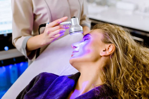 Skin Cosmetology. Woman beautician Doing Blue Light Therapy On face of pretty young blond woman