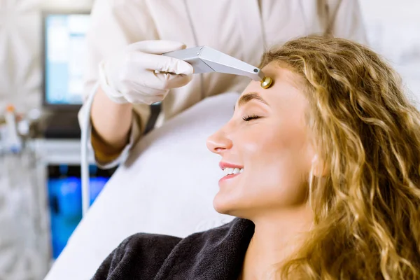 Microdermabrasion clinic treatment, aesthetic facial procedure, anti acne and hyperpigmentation cosmetic. Cosmetologist hand doing the microdermabrasion for young smiling blond woman.