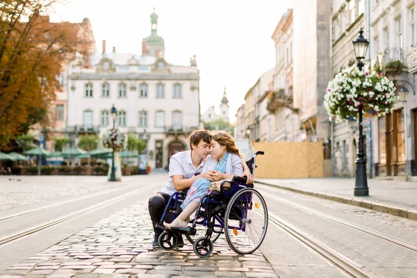 Verliebtes Paar in der Altstadt. hübsches junges Mädchen mit Krankheit im Rollstuhl und ihrem liebenswerten Mann, der geht, lächelt und die Stirn berührt, die Augen schließt — Stockfoto