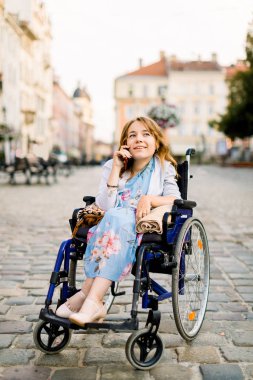 Tekerlekli sandalyedeki genç ve güzel bir kadının portresi. Günün tadını çıkarıyor ve dışarıda yürürken telefonla konuşuyor.