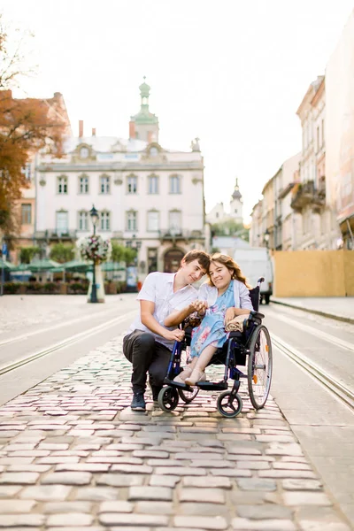 Молодий красивий усміхнений чоловік і його гарна дружина в інвалідному візку позує разом на тлі старого центру міста. Концепція кохання, інвалідності — стокове фото