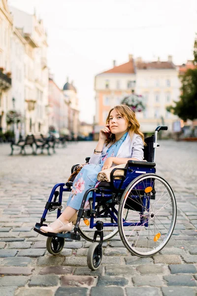 Behindertes junges Mädchen im Rollstuhl spricht auf dem Handy, im Freien, vor dem Hintergrund alter städtischer Gebäude — Stockfoto