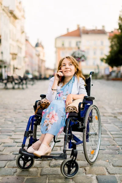 Porträt einer hinreißenden jungen blonden Frau im Rollstuhl, die ihren Tag genießt und telefoniert, während sie draußen in der Stadt spaziert — Stockfoto