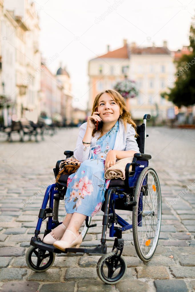 Portrait of a gorgeous young blond woman in a wheelchair enjoying her day and talking on the phone while walking in the city outdoors