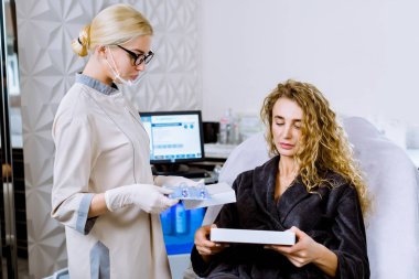 Genç sarışın kozmetik uzmanı, modern klinikteki kanepede oturan, genç sarışın, kıvırcık kadın müşteriyle kozmetik enjeksiyonları için yeni bir araç hakkında konuşuyor.