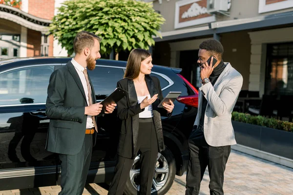 Tre unga multietniska affärsmän diskuterar något utomhus nära svart bil. Kaukasiska kvinnor har surfplatta, afrikansk man talar i telefon, och kaukasisk man med affärskontrakt — Stockfoto