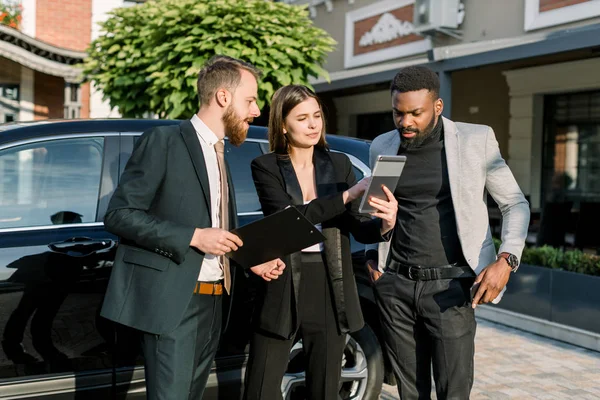 Two men, African and Caucasian, and one Caucasian woman stand in front of black car on a parking place on yard of dealer. Woman showing something on the digital tablet — 스톡 사진