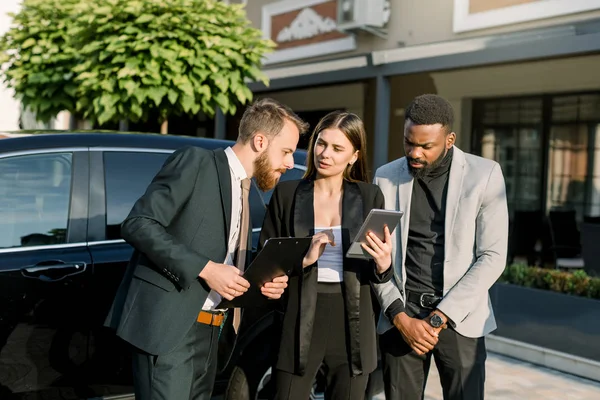 Briefing rapide avant la réunion. Trois jeunes gens d'affaires multiethniques joyeux, deux hommes et une femme, se parlent tout en se tenant à l'extérieur près de la voiture noire. Femme détient tablette numérique — Photo