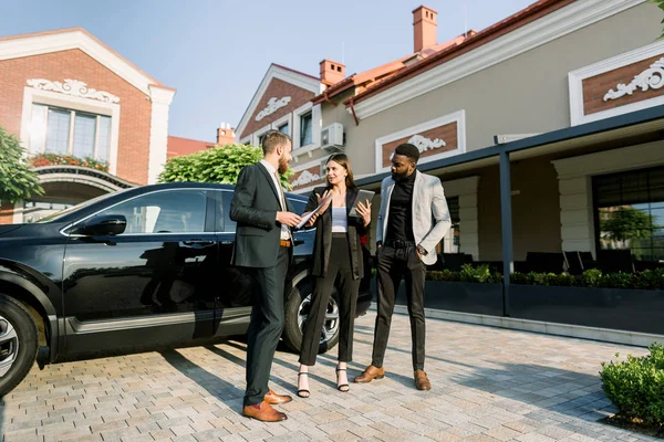 Penjual mobil laki-laki muda Kaukasia bekerja dengan klien, pasangan bisnis pria Afrika dan wanita Kaukasia, di dealer luar ruangan, menunjukkan crossover mobil baru hitam — Stok Foto