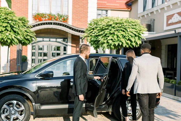 Chauffeur ouvrant porte de voiture pour couple d'affaires, femme blanche et homme africain, vue de côté. Voyage d'affaires en voiture . — Photo