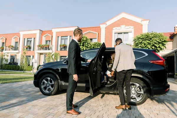 Jeune homme d'affaires caucasien en costume ouvrant porte de voiture noire pour ses collègues, homme africain et femme caucasienne. En plein air, bâtiments du centre d'affaires — Photo