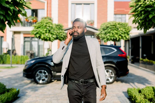 Elegante Afrikaanse zakenman wandelen in de straat, terwijl het nemen van aan de telefoon, zwarte auto crossover en bouwen op de achtergrond — Stockfoto