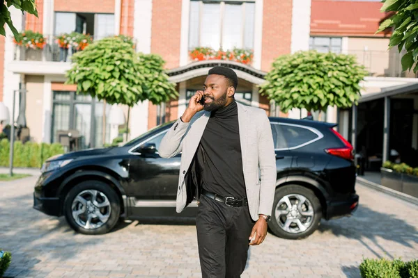 現代のビジネスセンターの庭や駐車場を歩いている間に電話で話してスマートなカジュアルなビジネススーツで深刻な豊かなアフリカのビジネスマン。背景に黒い車のクロスオーバー — ストック写真