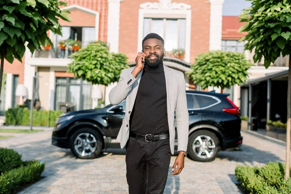 Jovem sorridente homem de negócios preto em um terno falando por smartphone andando ao ar livre perto de um carro preto e edifícios de tijolos modernos — Fotografia de Stock