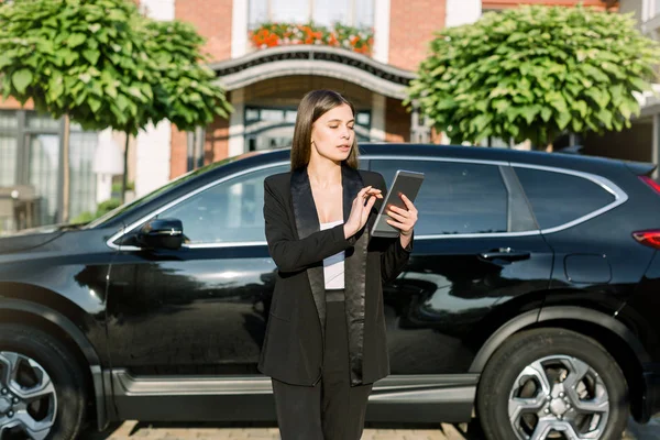 Young Caucasian businesswoman in black suit working on digital tablet outdoor standing near the black car over green trees and building background — 스톡 사진