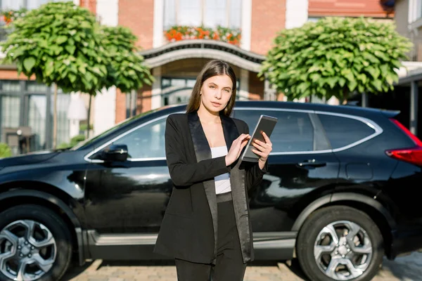 Young serious dark-haired business woman with tablet outdoors, standing in front of new black car crossover and modern brick ofiice building — 스톡 사진