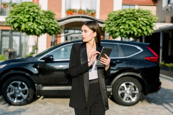 Manajer perempuan profesional, pengusaha Kaukasia dengan tablet yang berdiri di dekat mobil hitam di latar belakang halaman gedung kantor — Stok Foto