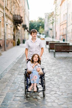 Tekerlekli sandalyedeki genç çift şehirde geziniyor. Tekerlekli sandalyedeki güzel genç kadın ve eski şehrin ortasında oturan erkek arkadaşı.