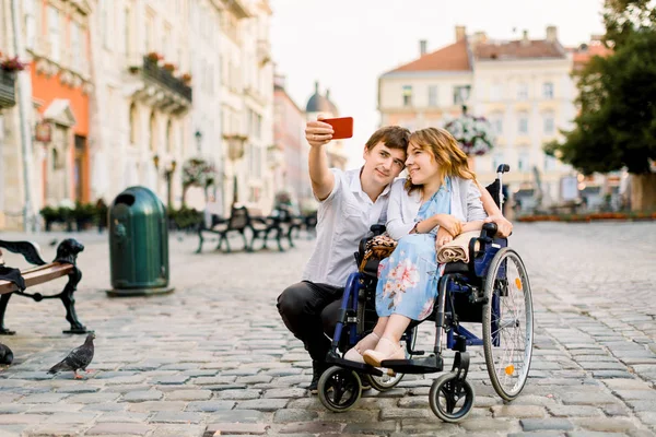 Selfie tijd. Knappe gelukkige jonge man knuffelen zijn gehandicapte jonge mooie vrouw en glimlachen terwijl het nemen van selfie met haar in de stad — Stockfoto