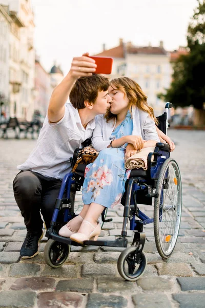 Junges Paar beim Selfie. ziemlich glückliche junge behinderte Frau im Rollstuhl küsst ihren jungen Freund und lächelt, während sie ein Selfie mit ihm in der Stadt macht — Stockfoto