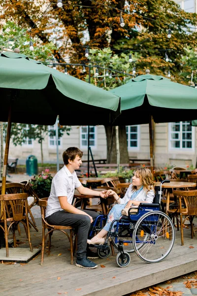 Молода красива жінка в інвалідному візку проводить час разом з чоловіком, тримаючись за руки, у відкритому кафе в старому центрі міста — стокове фото