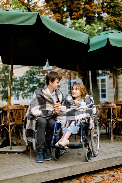 Zwei Freunde, schönes Paar Mann und Frau mit karierten Plaids in einem Café im Freien Kaffee trinken, der Mann sitzt im Rollstuhl — Stockfoto