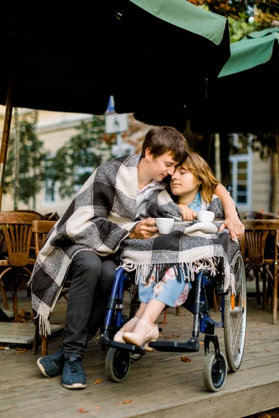 Schöner junger Mann, der an einem sonnigen Tag Kaffee trinkt und sich mit einer hübschen Frau im Rollstuhl trifft. Mann umarmt Frau in karierten Karos im Café — Stockfoto