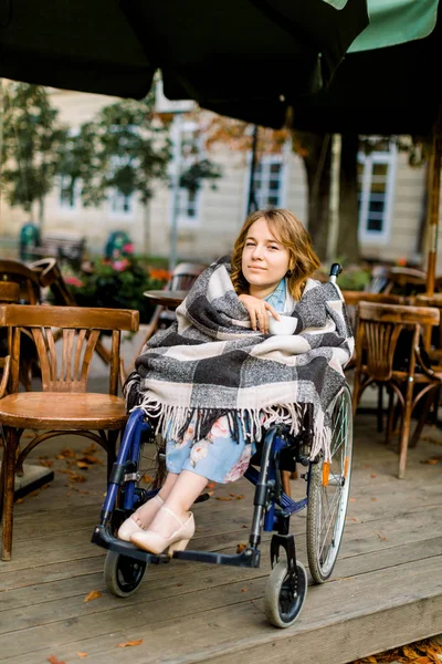 Πορτραίτο μιας νεαρής όμορφης γυναίκας σε αναπηρικό καροτσάκι, καλυμμένο με καρό καρό καρό, πίνοντας καφέ ενώ κάθεται σε εξωτερικό χώρο cafe. — Φωτογραφία Αρχείου