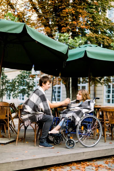 Porträt einer hübschen jungen Frau im Rollstuhl, die sich bei einem Date mit ihrem gutaussehenden Mann amüsiert, Kaffee trinkt und Händchen hält, in einem Café in der Stadt sitzt — Stockfoto