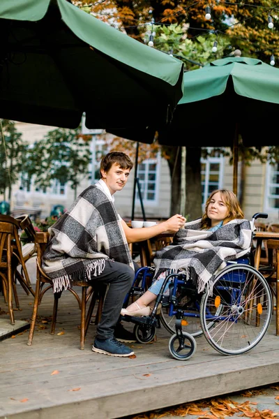 Porträt einer hübschen jungen Frau im Rollstuhl, die sich bei einem Date mit ihrem gutaussehenden Mann amüsiert, Kaffee trinkt und Händchen hält, in einem Café in der Stadt sitzt — Stockfoto