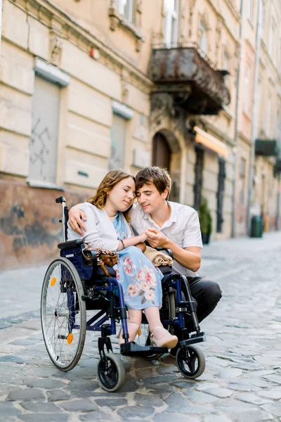 Ein junger Mann umarmt seine hübsche Frau im Rollstuhl, während er gemeinsam auf der Straße der Stadt spaziert. Verliebtes Paar im Rollstuhl umarmt und berührt Stirn mit geschlossenen Augen, draußen in der Stadt — Stockfoto
